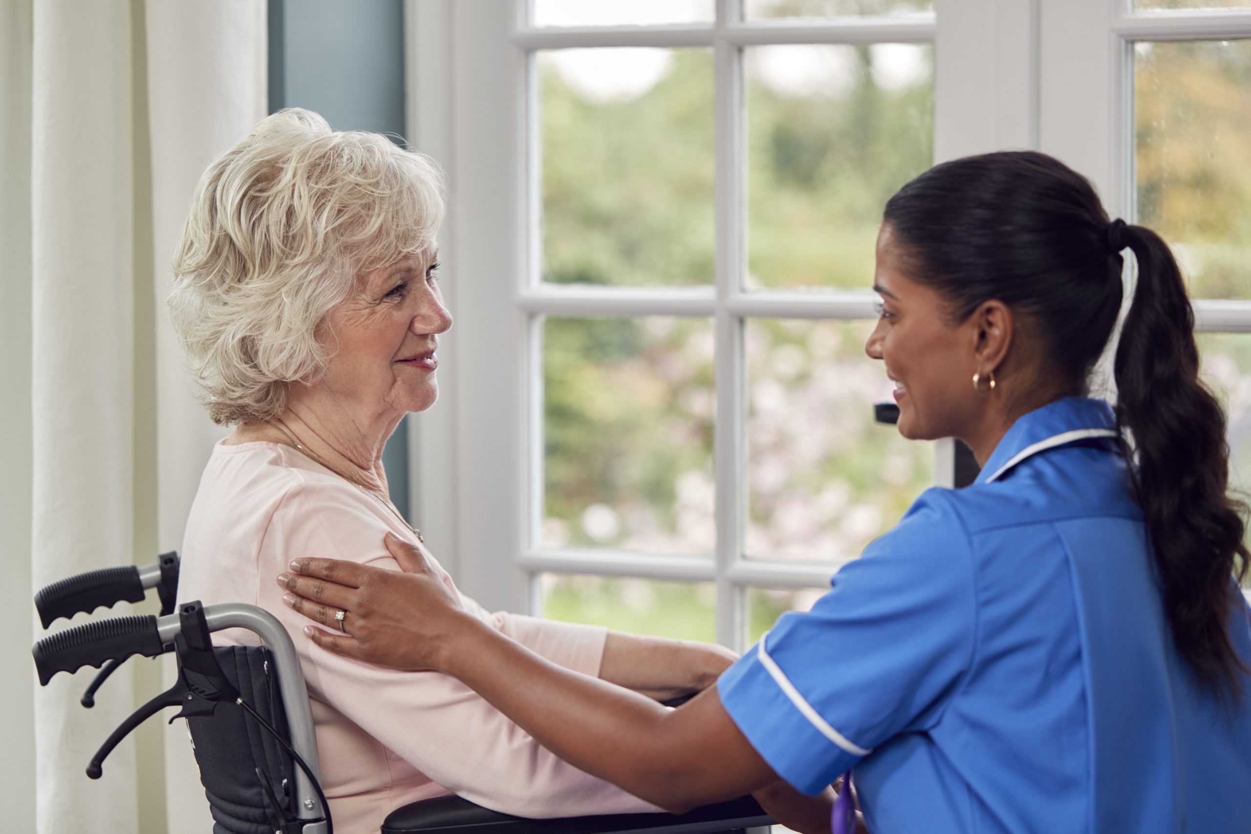 Image showcasing a female caregiver, dressed in a uniform, engaged in conversation with a senior woman seated in a wheelchair in the lounge of a care home.