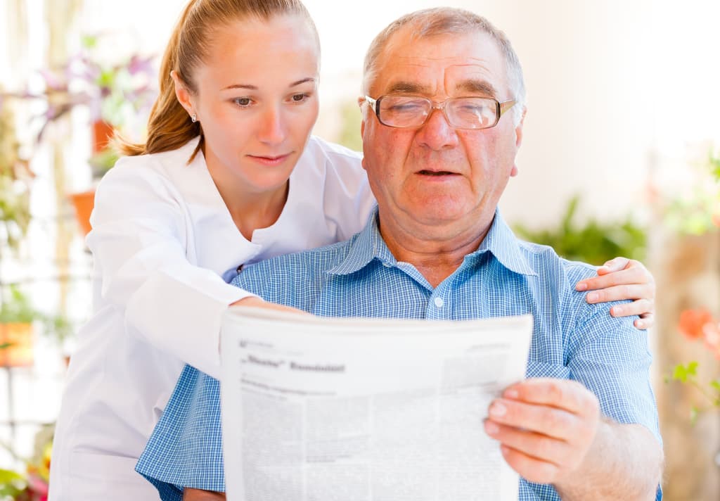 A woman reads a newspaper to an elderly man, sharing news and moments of connection in a warm, inviting setting.