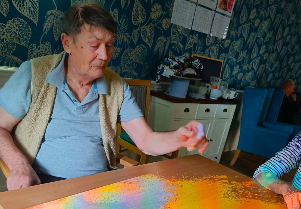An elderly man and a young girl are seated at a table, engaged in painting with various coloured paints.