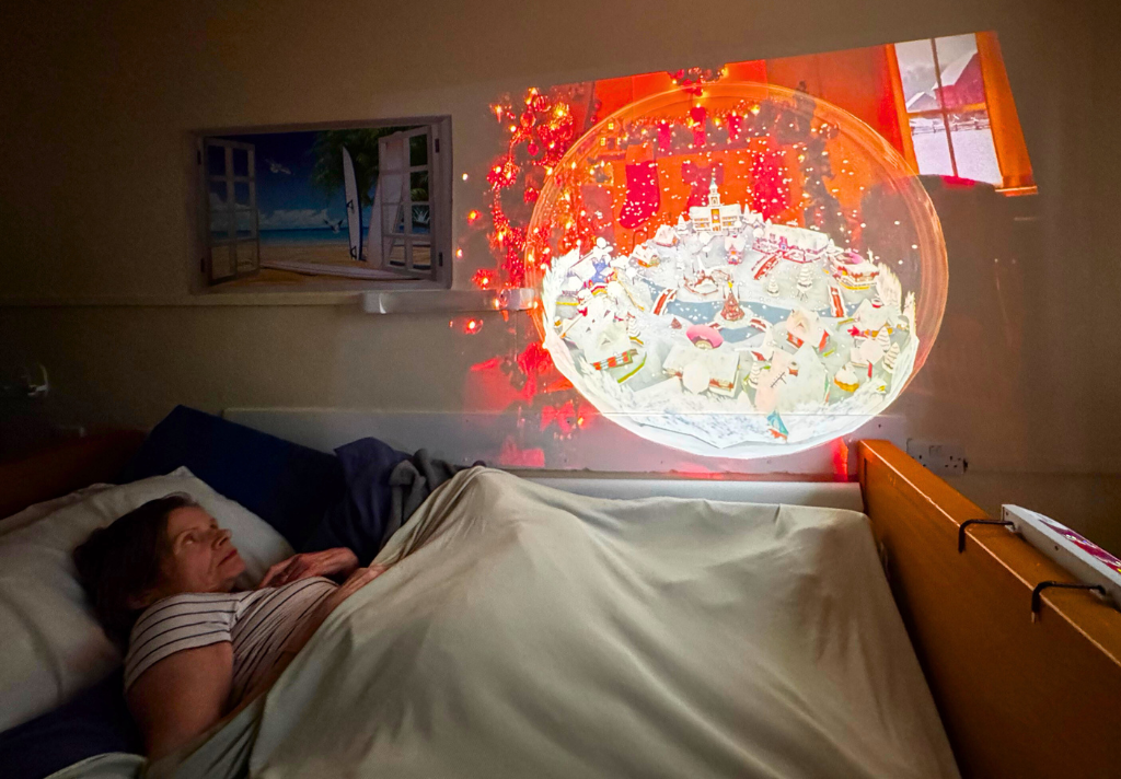 A woman lies in bed, gazing at a large projection of a Christmas tree on the wall above her.