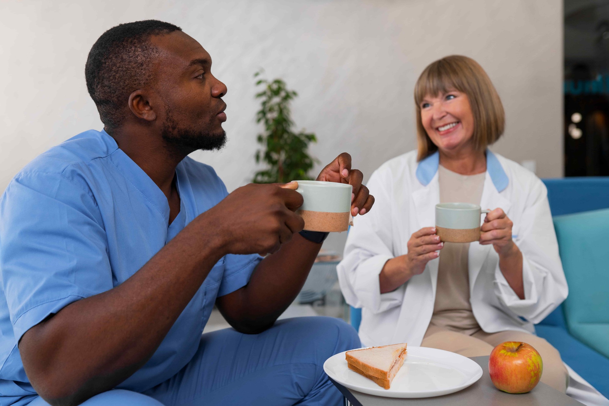 Care home staff member and senior colleague sharing a tea break and conversation in a comfortable lounge setting.