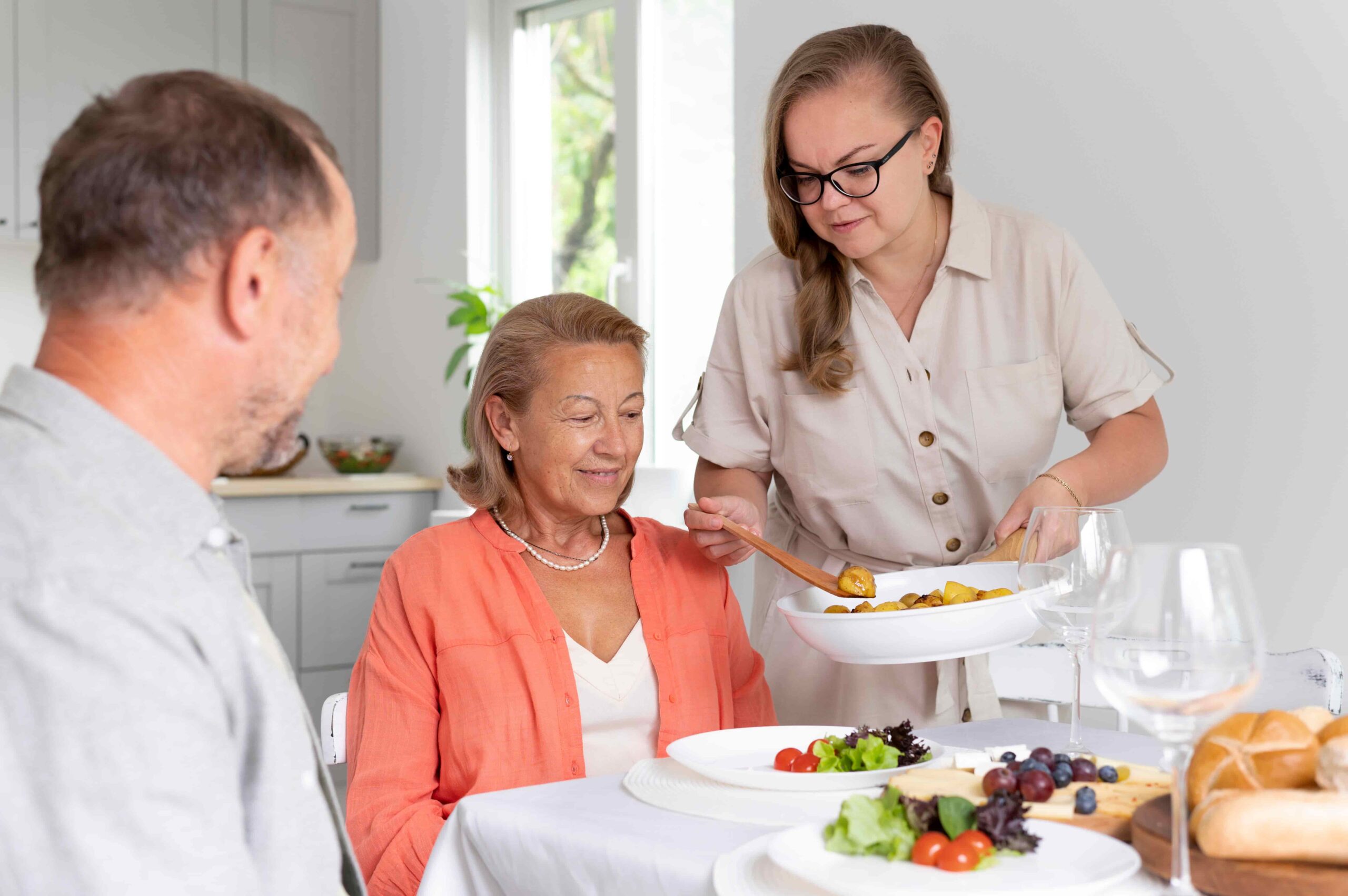 A person serving a dish of roasted potatoes at a dining table set with plates of salad, grapes, bread rolls, and wine glasses, while two other people sit at the table in a bright kitchen setting.