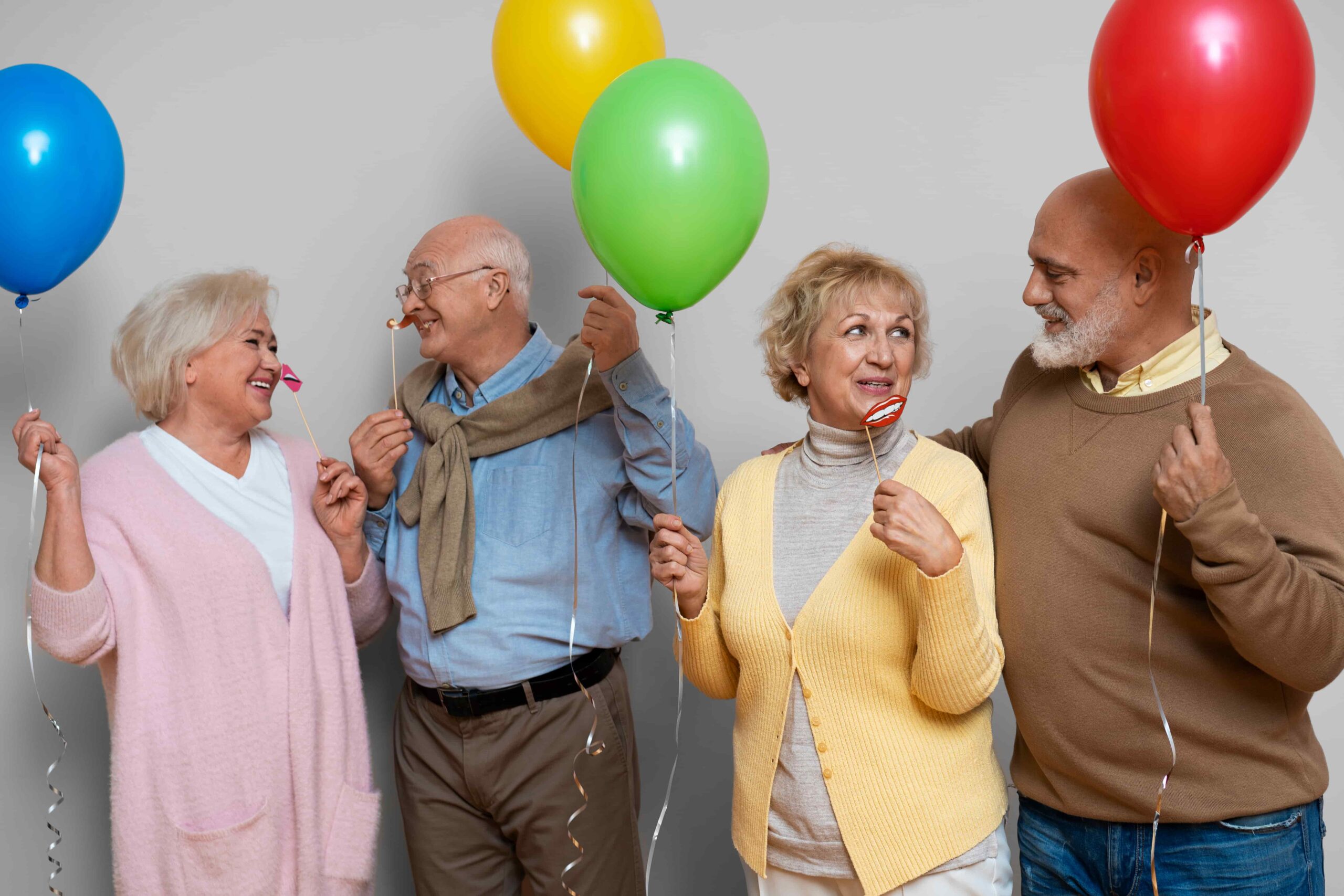 Four older adults standing together holding colorful balloons and party props against a plain background, dressed in casual sweaters and cardigans, appearing to celebrate at a festive gathering.
