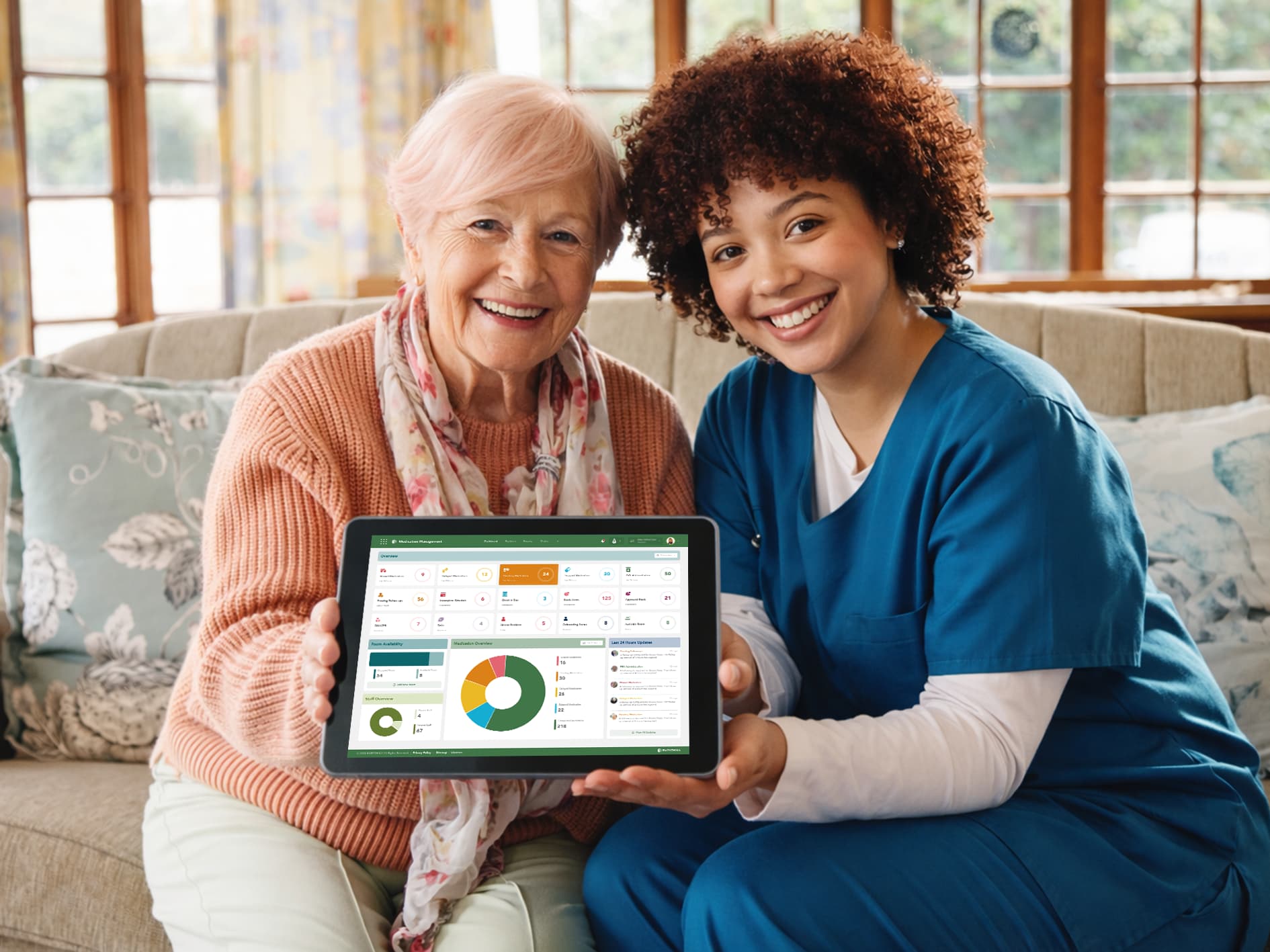 A caregiver and an older adult sitting together on a sofa, jointly holding a tablet displaying a colourful care‑management dashboard.