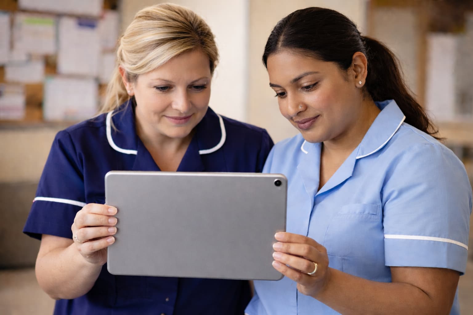 Two healthcare workers in uniform standing close together and reviewing information on a tablet device in a clinical setting.