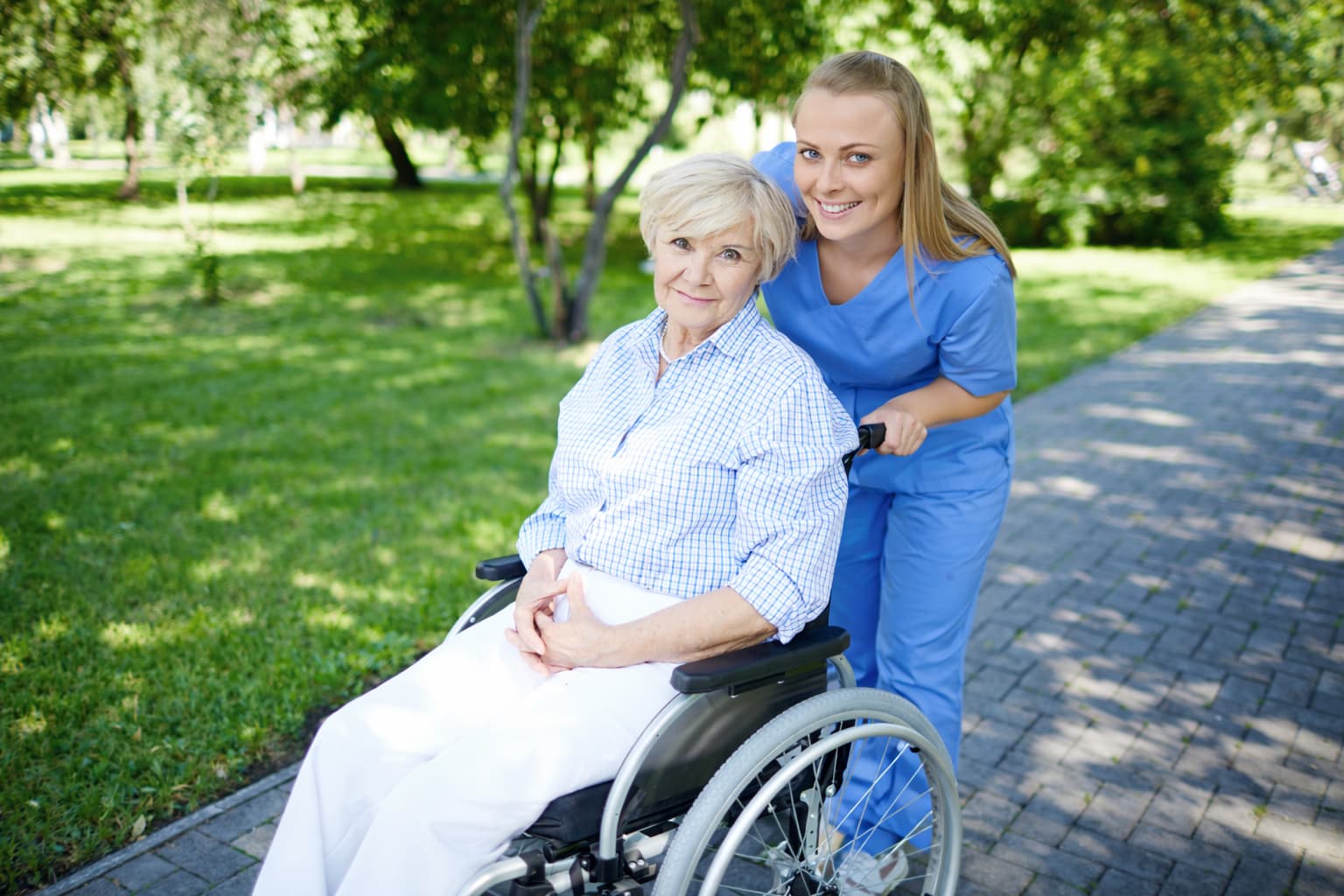 Caregiver in blue scrubs standing behind an elderly person in a wheelchair on a sunny park path.