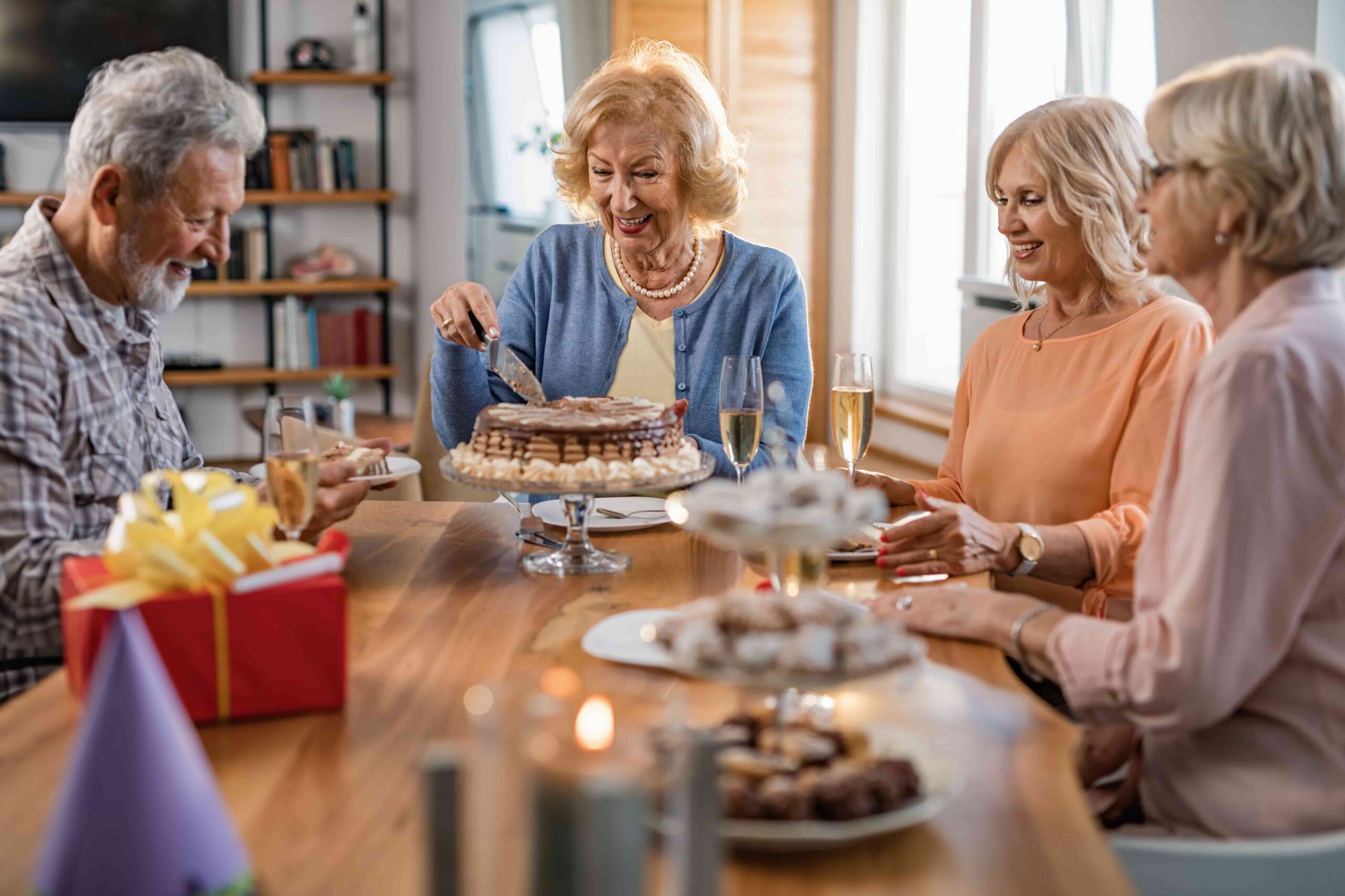Group of older adults celebrating around a cake and gifts at a dining table, smiling and enjoying champagne.