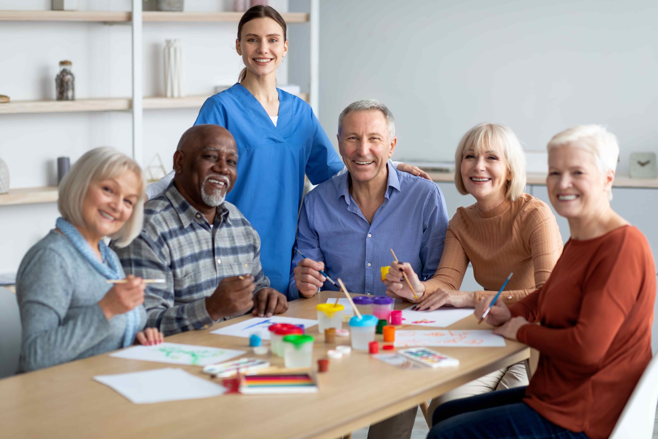 Cheerful nurse in blue scrubs posing with a group of smiling older residents during an art therapy session in a bright care home setting.