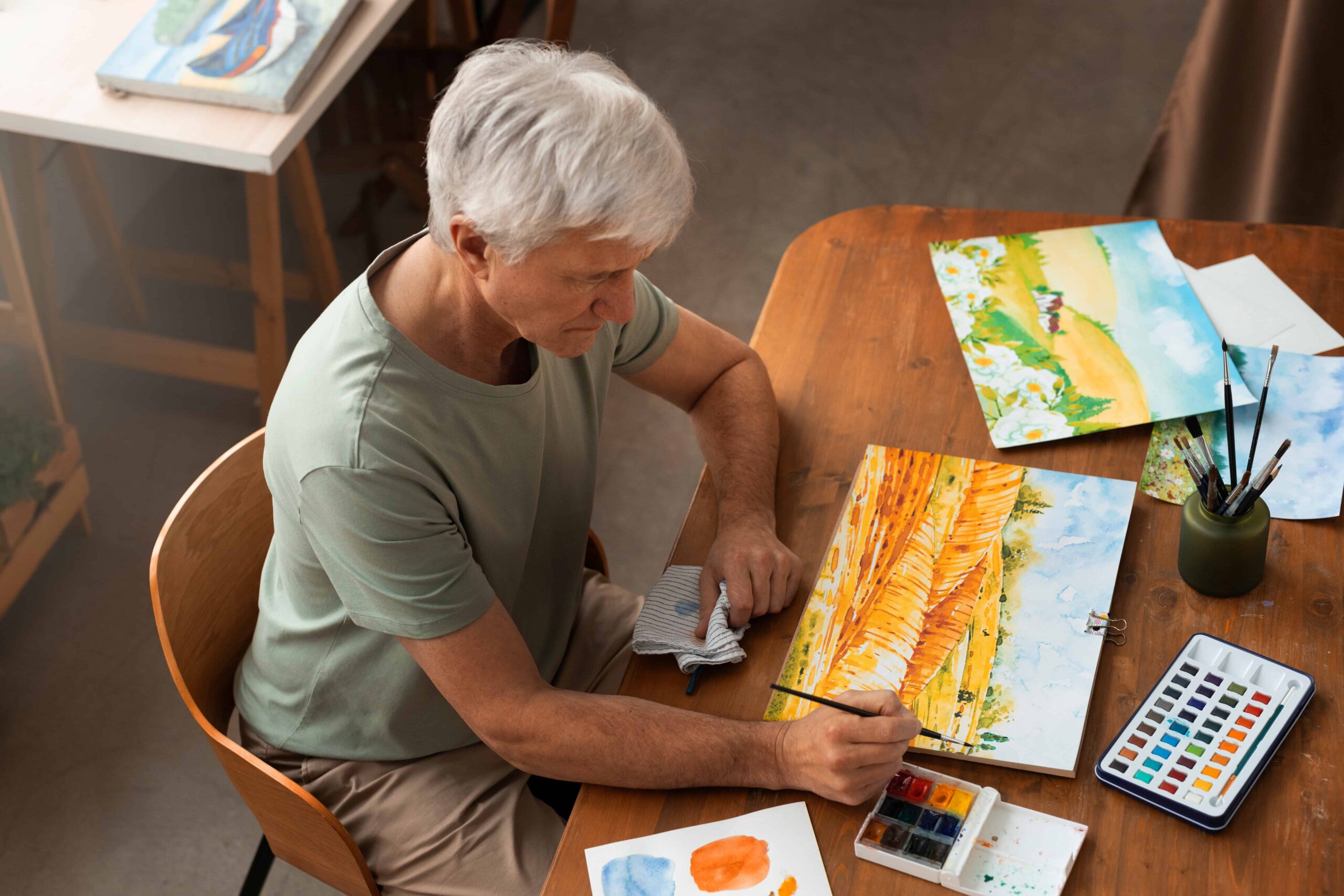 Person painting a vibrant watercolour landscape at a wooden table, surrounded by brushes, palettes, and other completed artwork.