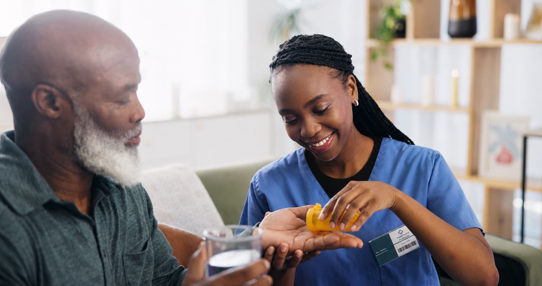 Healthcare worker in blue scrubs pouring medication into an older adult’s hand while the older adult holds a glass of water in a home setting.