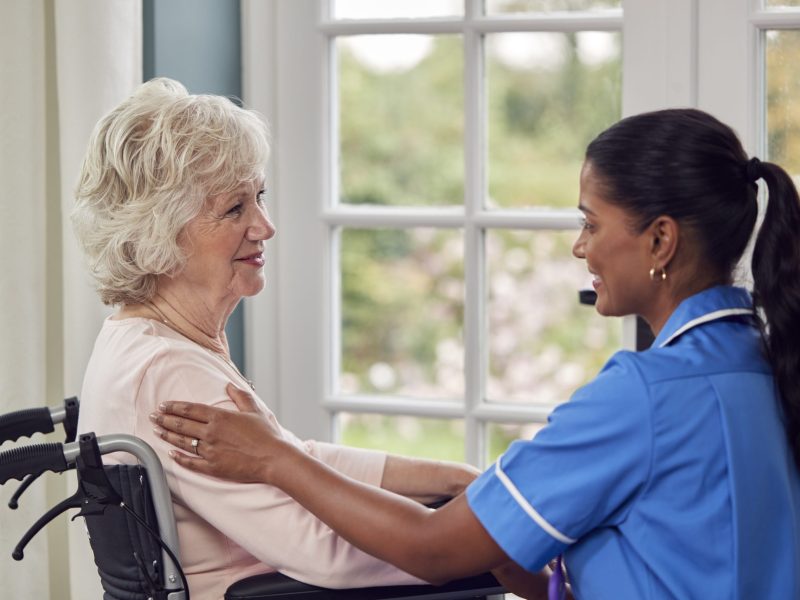 Image-showcasing-a-female-caregiver-dressed-in-a-uniform-engaged-in-conversation-with-a-senior-woman-seated-in-a-wheelchai-scaled.jpg